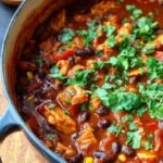 A steaming bowl of Mexican chicken chili topped with fresh avocado and cilantro in a dark ceramic bowl.