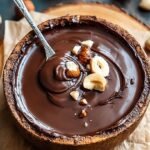 A glass jar filled with smooth, glossy homemade nutella next to a pile of roasted hazelnuts.