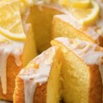 A golden, glazed Keto Lemon Pound Cake sliced on a wooden board with lemon slices.