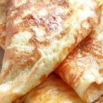 A stack of thin, golden low carb crepes served with fresh berries and whipped cream on a white plate.