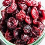 A bowl of vibrant red sugar free dried cranberries on a wooden table with fresh berries nearby.