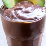 A thick chocolate avocado smoothie in a glass with a straw and fresh mint garnish