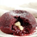 A warm red velvet lava cake in a white ramekin with a molten chocolate center flowing out.