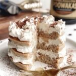 A stack of fluffy tiramisu protein pancakes layered with cream and dusted with cocoa powder on a white plate.