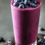 A creamy purple blueberry smoothie in a tall glass with a straw and fresh berries on top.
