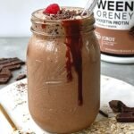 A tall glass of chocolate oatmeal protein smoothie with a dusting of cocoa powder on top.