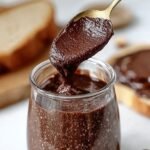 Smooth chocolate hazelnut spread in a glass jar with whole hazelnuts nearby