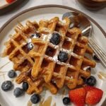 A stack of golden brown sweet potato protein waffles topped with maple syrup and pecans on a white plate.