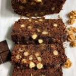 A sliced loaf of dark chocolate zucchini bread with melting white chocolate chips on a wooden board.