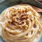 A warm bowl of trofie pasta coated in a creamy, pale walnut sauce topped with crushed nuts and fresh herbs.