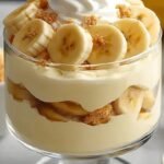 A glass trifle dish filled with layers of creamy vanilla pudding, sliced bananas, and vanilla wafer cookies topped with crushed crumbs.