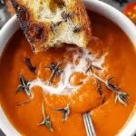 A bowl of creamy roasted tomato soup topped with fresh basil leaves and a side of grilled cheese.