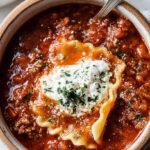 A bowl of rich red lasagna soup topped with a creamy white ricotta cheese dollop and fresh herbs