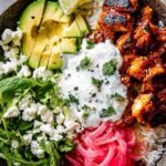 A vibrant grain bowl with glazed chicken, cucumber, tomatoes, and white sauce