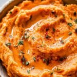 A bowl of smooth, orange sweet potato mash topped with a pat of melting butter and a drizzle of maple syrup