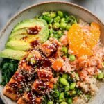 A colorful bowl of glazed salmon cubes, jasmine rice, sliced cucumbers, and julienned carrots topped with sesame seeds.