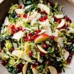 A bright bowl of shaved Brussels sprout salad topped with apple slices, pomegranate arils, and ribbons of Manchego cheese.