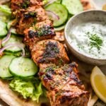 Grilled skewers of marinated chicken served with lemon and fresh herbs on a plate.