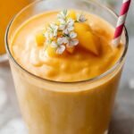 A tall glass of creamy orange mango lassi topped with a pinch of cardamom