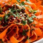 A bright orange grated carrot salad with green cilantro and red pepper flakes in a glass bowl.
