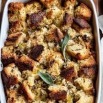 A golden brown sourdough stuffing in a white baking dish with fresh herbs on top.