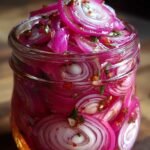 A glass jar filled with vibrant pink sliced red onions in a clear pickling liquid with red pepper flakes.