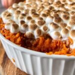 A golden-brown sweet potato casserole topped with toasted marshmallows and chopped pecans in a white baking dish.