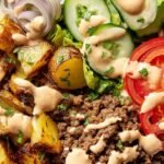 A colorful healthy burger bowl with browned beef, cherry tomatoes, avocado, and drizzled sauce.