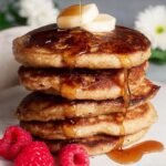 A stack of golden brown almond flour banana pancakes topped with fresh fruit and syrup.