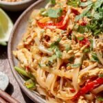 A steaming bowl of Healthy Chicken Pad Thai topped with peanuts, cilantro, and fresh lime wedges.