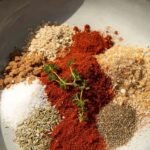 A small glass bowl filled with a colorful blend of ground turkey seasoning spices next to a skillet of browned meat.