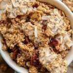 A close-up of golden brown chunky coconut granola with large clusters and toasted pecans on a baking sheet.