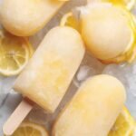A tray of bright yellow frozen lemonade popsicles with fresh lemon slices on a wooden table.