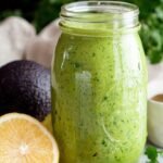 A bowl of vibrant green avocado dressing surrounded by fresh herbs and lemon slices.