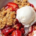 A golden brown strawberry crumble in a white baking dish with fresh halved strawberries