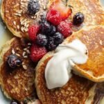 A stack of golden oat flour pancakes topped with fresh berries and maple syrup on a white plate.