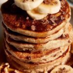 A stack of golden brown banana pancakes topped with walnuts and chocolate chips