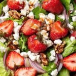 A fresh spinach salad topped with sliced strawberries, crumbled goat cheese, and toasted walnuts in a large bowl.