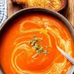 A bowl of smooth, orange-red tomato soup swirled with coconut milk and topped with fresh basil.