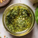 Fresh green walnut pesto in a glass jar surrounded by basil leaves and walnuts