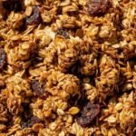 A golden tray of homemade granola with nuts, seeds, and dried cranberries on parchment paper.