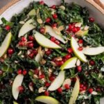 A bright green kale salad topped with red pomegranate arils, white feta, and toasted walnuts in a wooden bowl.