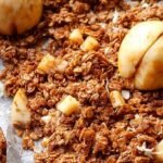 A close-up of crunchy granola with pecans and dried apple pieces on a baking sheet.