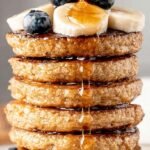A stack of golden brown protein oatmeal blender pancakes topped with fresh banana slices and maple syrup.