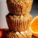 A stack of golden brown carrot orange muffins on a wire cooling rack with fresh orange slices nearby.