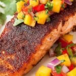 A seared blackened salmon fillet topped with colorful mango salsa on a white plate.