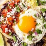 A plate of two crispy corn tortillas topped with black beans, fried eggs, red tomato sauce, and avocado slices.
