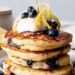 A tall stack of fluffy blueberry lemon pancakes topped with fresh berries and syrup
