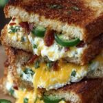 A golden brown grilled cheese sandwich sliced in half with melted cheese, roasted jalapenos, and bacon spilling out