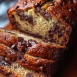 A golden loaf of chocolate chip banana bread sliced on a wooden board with melted chocolate chips visible.